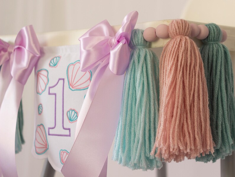White embroidered mermaid high chair banner with seashells, number one, and lavender bows for a girl’s first birthday.