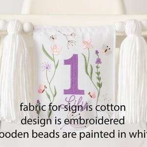 Pu&ograve; includere: Striscione in tessuto bianco con ricamo floreale, un grande numero 1 viola e il nome "Lily". Lo striscione &egrave; adornato con nappe bianche e perline di legno. &Egrave; visibile anche il testo "fabric for sign is cotton design is embroidered wooden beads are painted in white".