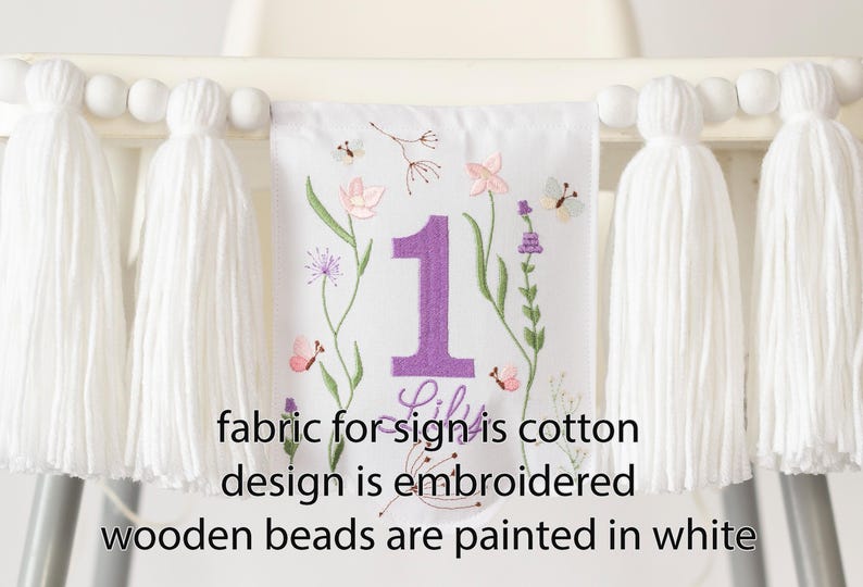 Pu&ograve; includere: Striscione in tessuto bianco con ricamo floreale, un grande numero 1 viola e il nome "Lily". Lo striscione &egrave; adornato con nappe bianche e perline di legno. &Egrave; visibile anche il testo "fabric for sign is cotton design is embroidered wooden beads are painted in white".