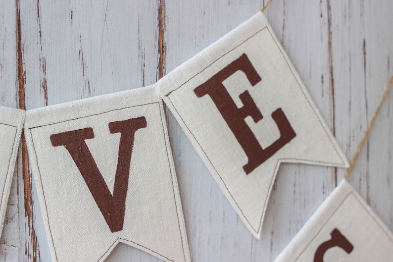 May include: Close-up of a fabric banner with the letters "V" and "E" in brown on a white background. The banner is strung with twine.