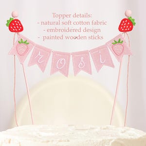 Embroidered name cake topper on pink cotton flags with strawberries on each end. Personalized topper for berry first birthday celebration.