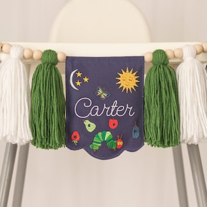 May include: A navy blue high chair banner with green and white tassels and wooden beads. The banner has the name "Carter" embroidered on it, along with a sun, moon, stars, butterfly, and caterpillar.
