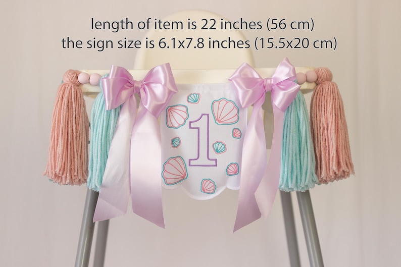 White embroidered mermaid high chair banner with seashells, number one, and lavender bows for a girl’s first birthday.