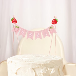 Embroidered name cake topper on pink cotton flags with strawberries on each end. Personalized topper for berry first birthday celebration.