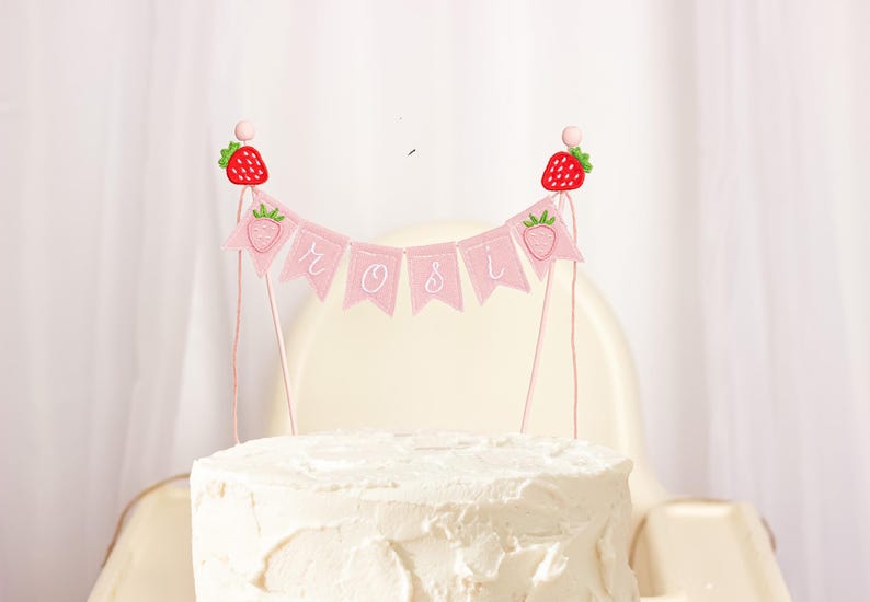 Embroidered name cake topper on pink cotton flags with strawberries on each end. Personalized topper for berry first birthday celebration.