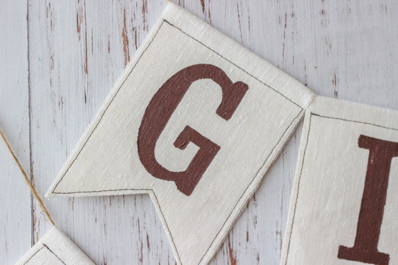 May include: Close-up of a brown fabric banner with the letters "G" and "I" painted in brown. The banner is sewn together with white thread.
