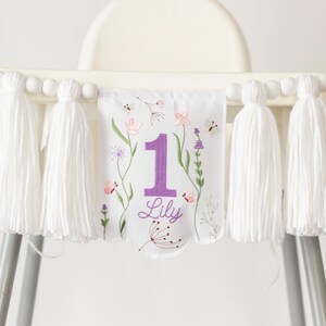 Pu&ograve; includere: Striscione per seggiolone bianco con grandi nappe bianche e perline di legno. Lo striscione presenta un motivo floreale con il numero 1 viola e il nome "Lily".