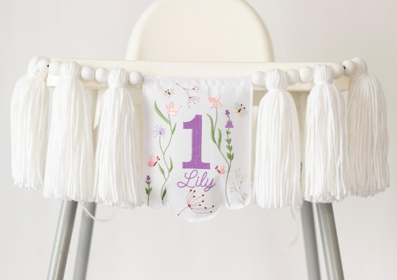 Pu&ograve; includere: Striscione per seggiolone bianco con grandi nappe bianche e perline di legno. Lo striscione presenta un motivo floreale con il numero 1 viola e il nome "Lily".