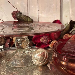 May include: A clear glass cake stand with a textured pedestal base, positioned next to a round, metallic silver ornament with white stripes and a red glass bowl featuring an etched grape pattern, all resting on a silver serving tray.