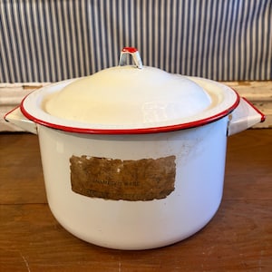 May include: White enamelware pot with a lid, featuring a red trim and handle. The pot has a label that reads "ENAMELED WARE". The pot has two side handles. The lid has a red handle. The pot is sitting on a wooden surface.