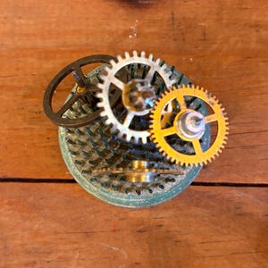 May include: A steampunk-style assemblage of watch gears and components. The piece features a central, textured, circular base in a teal-green hue, with various metal gears in silver and gold tones. The gears are arranged to create a unique, industrial-inspired design.