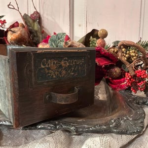 May include: A rectangular storage bin with a galvanized metal body and a dark wood front panel featuring a metal handle and a label plate with the text Cap Screws 1/4 x 2. The bin is positioned on a silver-toned tray with decorative holiday greenery.