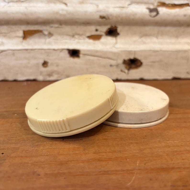 May include: Two off-white, oval-shaped lids. One lid is slightly raised, revealing a ridged edge. The other lid is flat. Both lids are on a wooden surface, with a distressed white background.