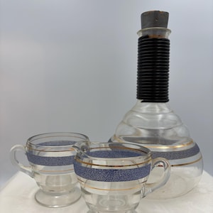 May include: A clear glass decanter with a black ribbed neck and cork stopper, accompanied by a creamer and a cup. Each piece features a blue speckled band with gold trim. The set is likely for serving beverages.