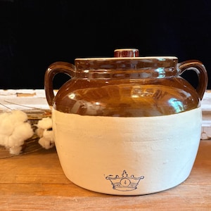 May include: A two-handled stoneware crock with a brown glazed upper section and a cream-colored lower section. The vessel features a matching lid and a blue stamped crown logo with the number 4 centered on the front of the cream base.