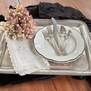 May include: A rectangular silver-plated serving tray with ornate handles, featuring a white ceramic plate with a dark rim, a set of silver flatware including two forks, a knife, and two spoons, a white lace-trimmed napkin, and dried hydrangea flowers.