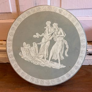 May include: A round, light blue decorative tin with a white relief design. The design features classical figures and a border of repeating floral patterns. The tin is likely vintage and suitable for storage or display.