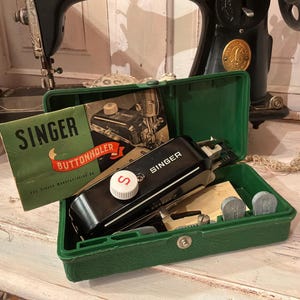 May include: A vintage Singer buttonholer attachment housed in a green plastic case. The set includes a black metal buttonholer with a white dial marked with an S, a green instruction manual titled SINGER BUTTONHOLER, and several metal cams.