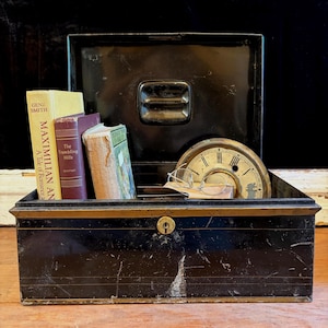 May include: An open, antique black metal box with a handle, containing several vintage books, a clock face, and eyeglasses. The box has a gold trim and a keyhole. The books include titles like "Maximilian An" and "The Trembling Hills."