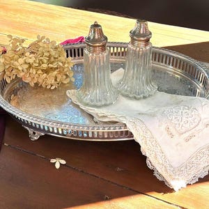 May include: An oval silver-plated serving tray with ornate handles and a pierced gallery rim, holding two clear glass salt and pepper shakers with silver-toned metal lids, a white lace-trimmed linen napkin, and dried hydrangea flowers.