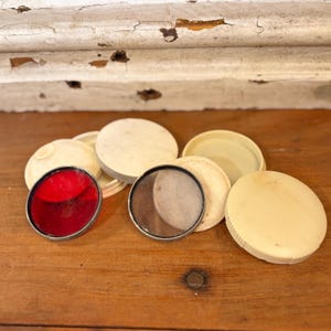 May include: A collection of vintage, round, cream-colored compacts and lids. One compact features a vibrant red filter, while another has a clear lens. The items are arranged on a wooden surface, suggesting a retro aesthetic.