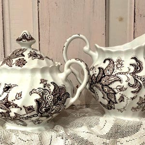 May include: A white ceramic sugar bowl with a lid and a matching creamer pitcher, both featuring a dark brown intricate floral and paisley pattern. The pieces have a scalloped rim, curved handles, and a fluted body design on a white base.