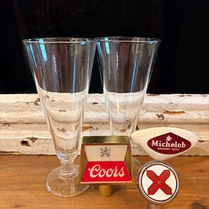 May include: Two clear, stemmed glasses and three vintage beer tap handles. The tap handles include a Coors handle with red and gold accents, a Michelob handle, and a handle with a red X design.