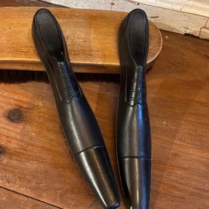 May include: Two black shoe brushes with handles shaped like formal dress shoes. The handles feature molded details representing laces and a shoe silhouette, while the brush bristles are positioned underneath the elongated, tapered black plastic bodies.