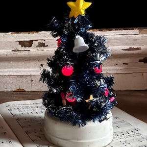 May include: A miniature artificial Christmas tree with dark blue tinsel needles, decorated with small red spherical ornaments, a white bell, and a yellow star topper. The tree is mounted on a plain white circular base.