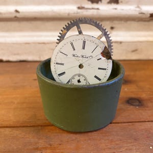 May include: An antique watch face with Roman numerals and the text "Western Watch Co" is set inside a small, olive green container. A metal gear is positioned behind the watch face. The items rest on a wooden surface.