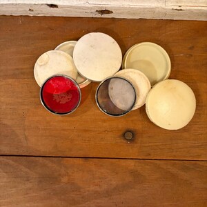 May include: A collection of vintage, round, cream-colored compacts and lids are arranged on a wooden surface. Two compacts have colored inserts: one red and one with a mesh screen. The lids vary in color and texture.