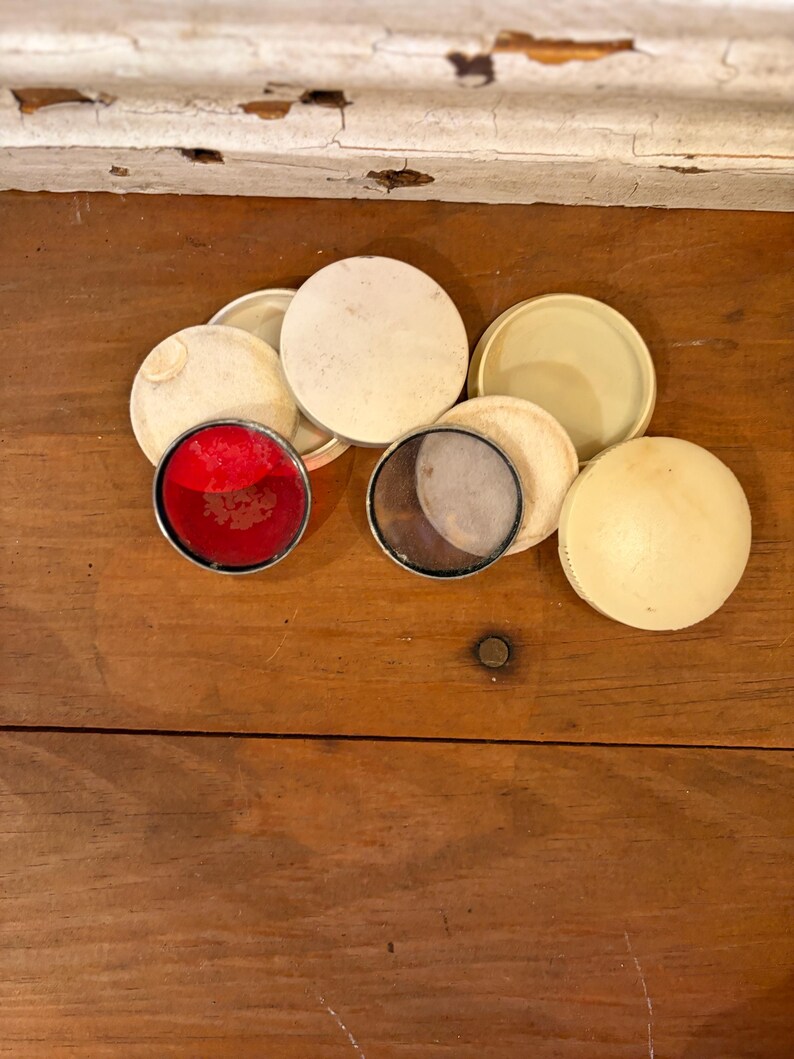 May include: A collection of vintage, round, cream-colored compacts and lids are arranged on a wooden surface. Two compacts have colored inserts: one red and one with a mesh screen. The lids vary in color and texture.
