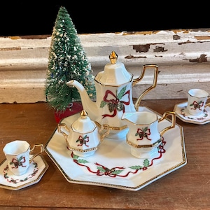 May include: A white porcelain tea set featuring a gold-trimmed hexagonal design with red ribbons, green holly leaves, and pinecone motifs. The set includes a teapot, a lidded sugar bowl, a creamer, two cups with saucers, and a large serving tray.
