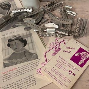 May include: A collection of silver-toned metal hair rollers and clips scattered alongside vintage instructional pamphlets featuring black and white photographs of hairstyles and text including LaMarick Special and Pretty Pageboy.