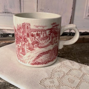 May include: A cream-colored ceramic mug featuring a red transferware print of a village scene with a church, houses, and trees. The mug has a visible crackle glaze finish, a cylindrical body, and a curved handle on the right side.