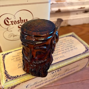 May include: A brown glass pipe with a carved face design, resting on a vintage Avon box. The box is cream-colored with gold accents and text. A tin with the text "Crosby Pipes" is in the background.