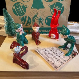 May include: A collection of five ceramic elf figurines in red and green outfits, including one doing a handstand, alongside a small green bottle brush tree and a piece of sheet music with printed lyrics, all displayed on a light-colored surface.