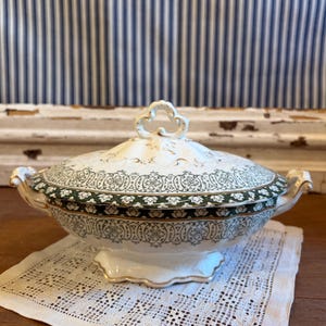 May include: A white and green ceramic serving dish with a lid. The dish features a decorative green and gold pattern around the rim and on the lid. The handles and lid finial are gold-colored.