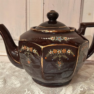 May include: A dark brown hexagonal teapot with a matching lid, featuring gold-painted trim along the edges and a delicate hand-painted floral pattern in orange, blue, and white. The teapot has a curved spout and a sturdy handle, with a glossy finish.