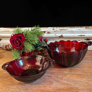 May include: Two deep red glass bowls with scalloped edges and integrated side handles. The bowls feature a vertical ribbed pattern along the exterior walls and a smooth, glossy finish. The glass is translucent, allowing light to pass through.