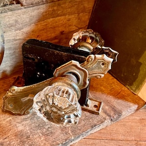 May include: A vintage door hardware set featuring two clear, faceted glass knobs attached to a black metal mortise lock mechanism. The assembly includes ornate, aged brass-toned metal backplates with decorative edges and mounting screw holes.