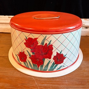 May include: A vintage cake carrier with a bright red lid and a white base. The sides are decorated with a red and green plaid pattern and red floral designs. A silver handle is on top.