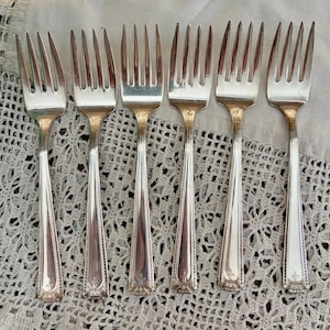 May include: A set of six silver-toned metal forks arranged in a row. Each fork features four tines and a slender handle with a decorative border pattern and a small engraved emblem at the base of the handle, showcasing a polished metallic finish.