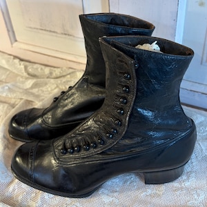 May include: A pair of antique black leather boots with button closures and rounded toes. The boots have a mid-calf height and a small heel. The leather appears worn, suggesting age and use. The boots are displayed on a lace-covered surface.