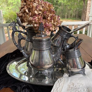 May include: A silver-plated tea service set on an oval tray, including a teapot, a creamer, and a lidded vessel. Each piece features ornate embossed floral bands, decorative finials, and tripod feet. Dried hydrangea flowers are tucked inside.