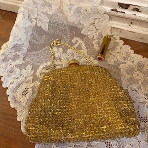 May include: A vintage gold beaded handbag with a gold chain strap and a gold lipstick case with a red lipstick. The handbag is resting on a white lace doily.