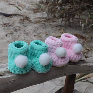 May include: Two pairs of baby booties, one pair is pink and the other is teal. Both pairs have white pom-poms.