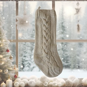 May include: A cream-colored, cable-knit Christmas stocking hangs in front of a window with a snowy winter scene. The stocking features a prominent cable-knit design down the center. The background includes a decorated Christmas tree and white ornaments.