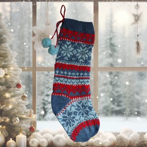 Puede incluir: Un calcetín de Navidad tejido en azul, rojo y azul claro, con un diseño de copo de nieve. Tiene un lazo rojo para colgar y está adornado con dos pequeños pompones azules. El fondo muestra un árbol de Navidad y una ventana.