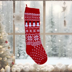 以下が含まれることがあります： 白い雪の結晶と雪だるまの模様が入った赤いクリスマスストッキング。編み物のストッキングには吊り下げ用のループがあり、雪景色のある窓の前に飾られています。